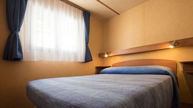 A hotel room with a bed, a window with curtains, and a bedside table with a lamp. - Camping Rio Martino  (Latina)