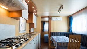 A compact kitchenette with a gas stove, sink, and white cabinets. There is a dining area with a table and chairs, and a window with curtains. - Camping Rio Martino  (Latina)