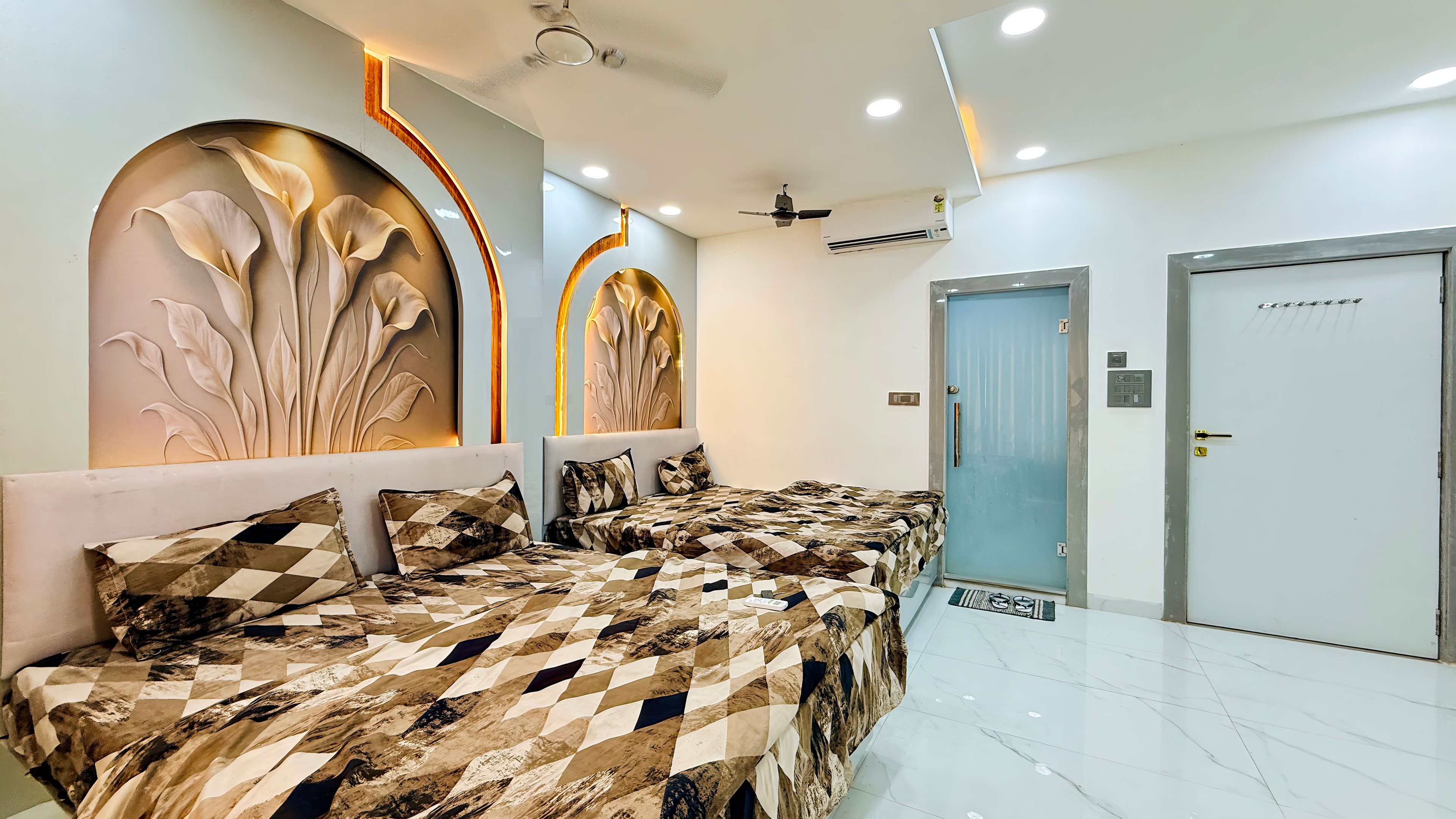A hotel room with a modern design, featuring a large bed with a patterned bedspread, a wall with decorative panels, a ceiling fan, and a door with a security lock.