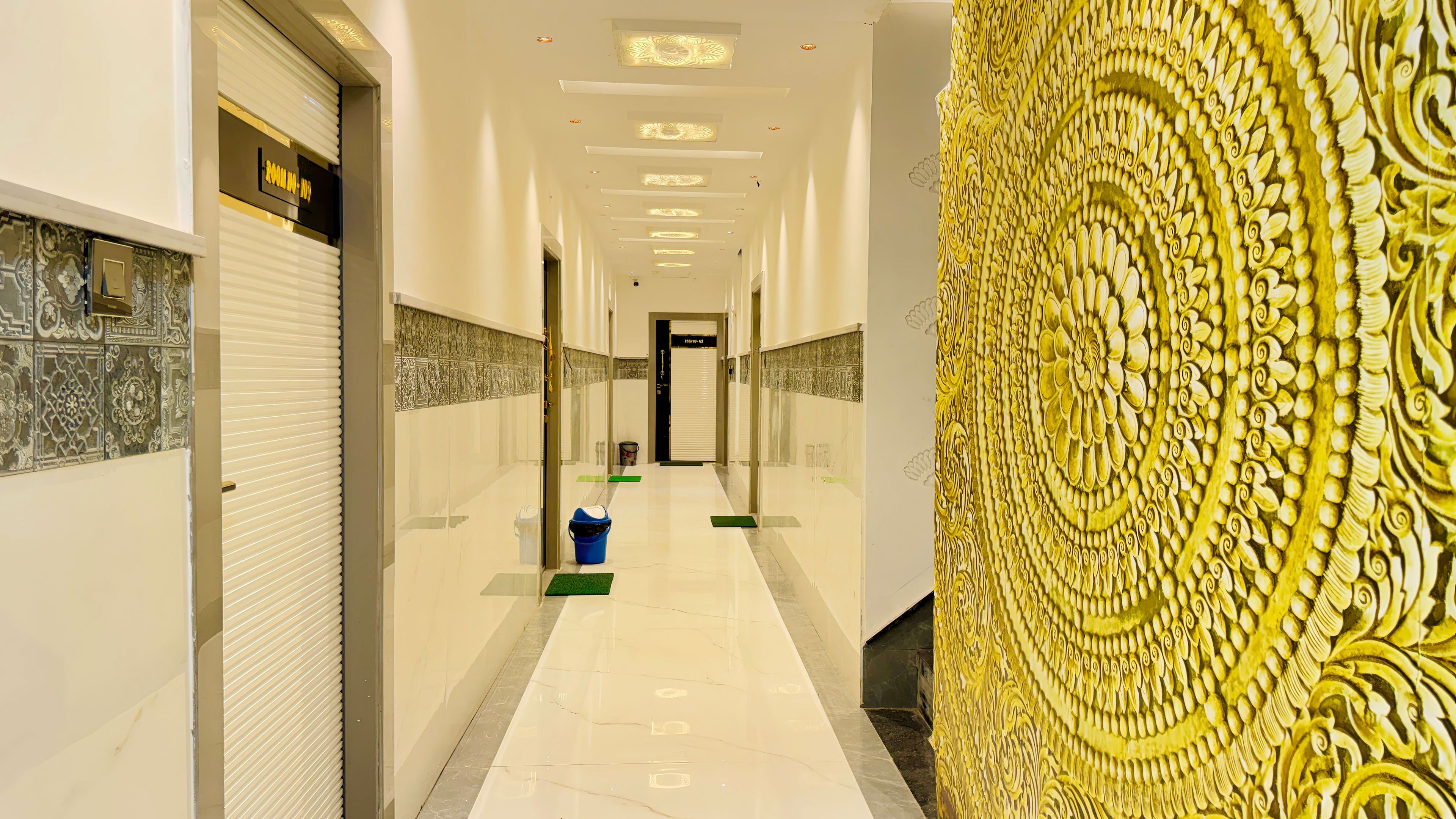 Corredor de hotel com piso ladrilhado, porta fechada à esquerda e parede decorativa à direita.