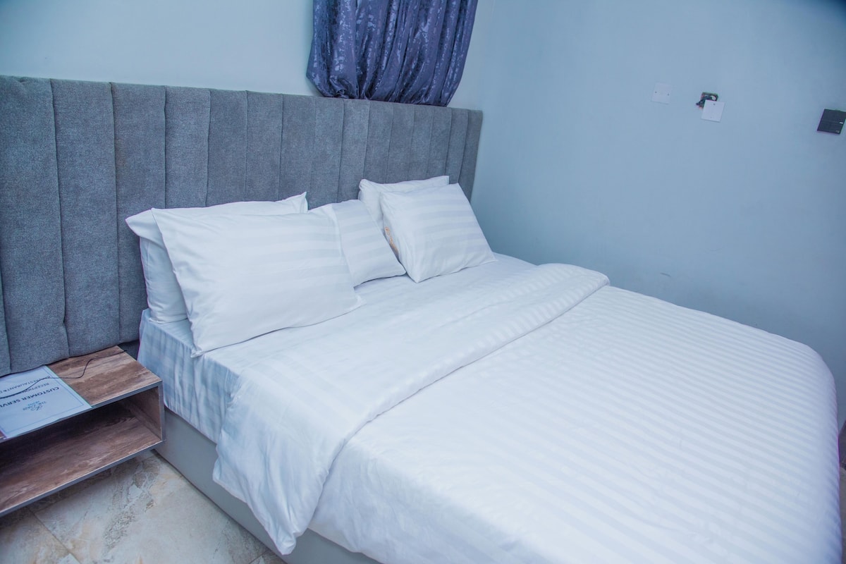 A neatly made bed with white linens, a grey upholstered headboard, and a wooden nightstand with a book.