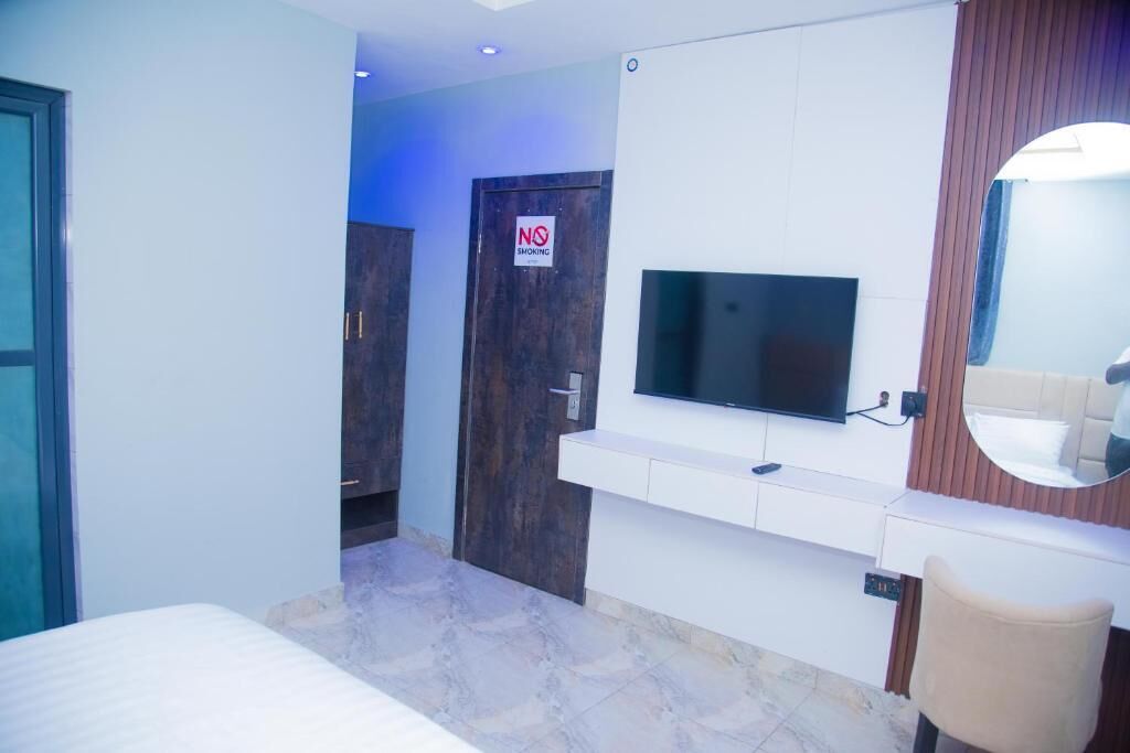 A modern hotel room with a bed, a TV mounted on the wall, a mirror, and a 'No Smoking' sign on the door.