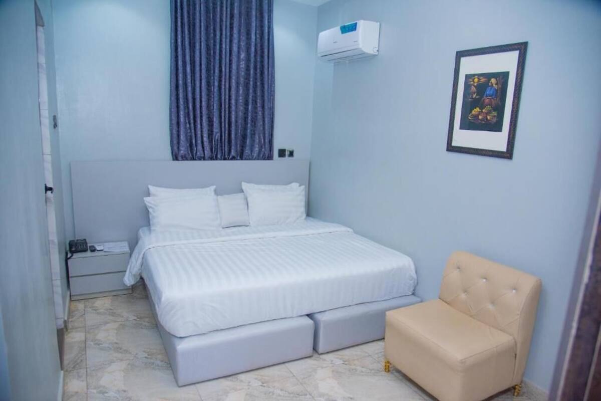 A bedroom with a bed, a bedside table, a chair, a wall-mounted air conditioner, and a framed picture.