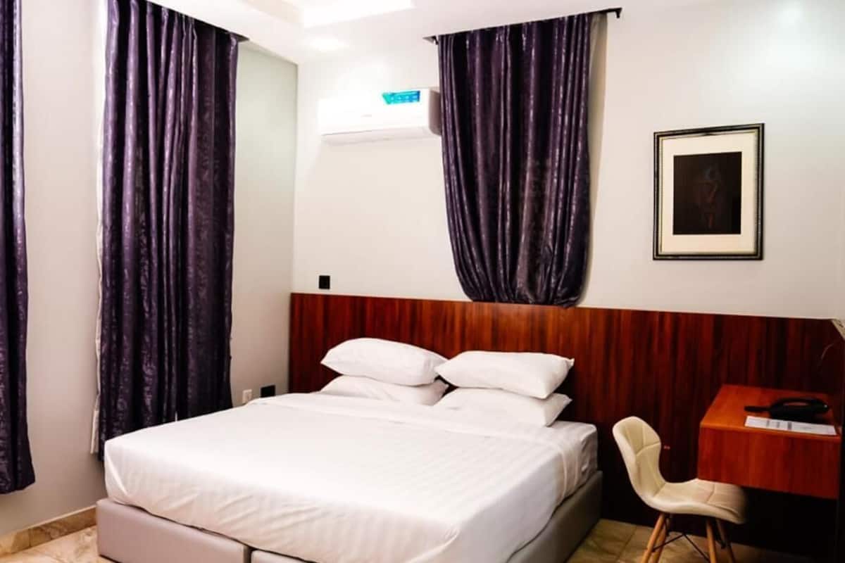 A hotel room with a bed, a wooden headboard, a desk with a chair, and a framed picture on the wall.