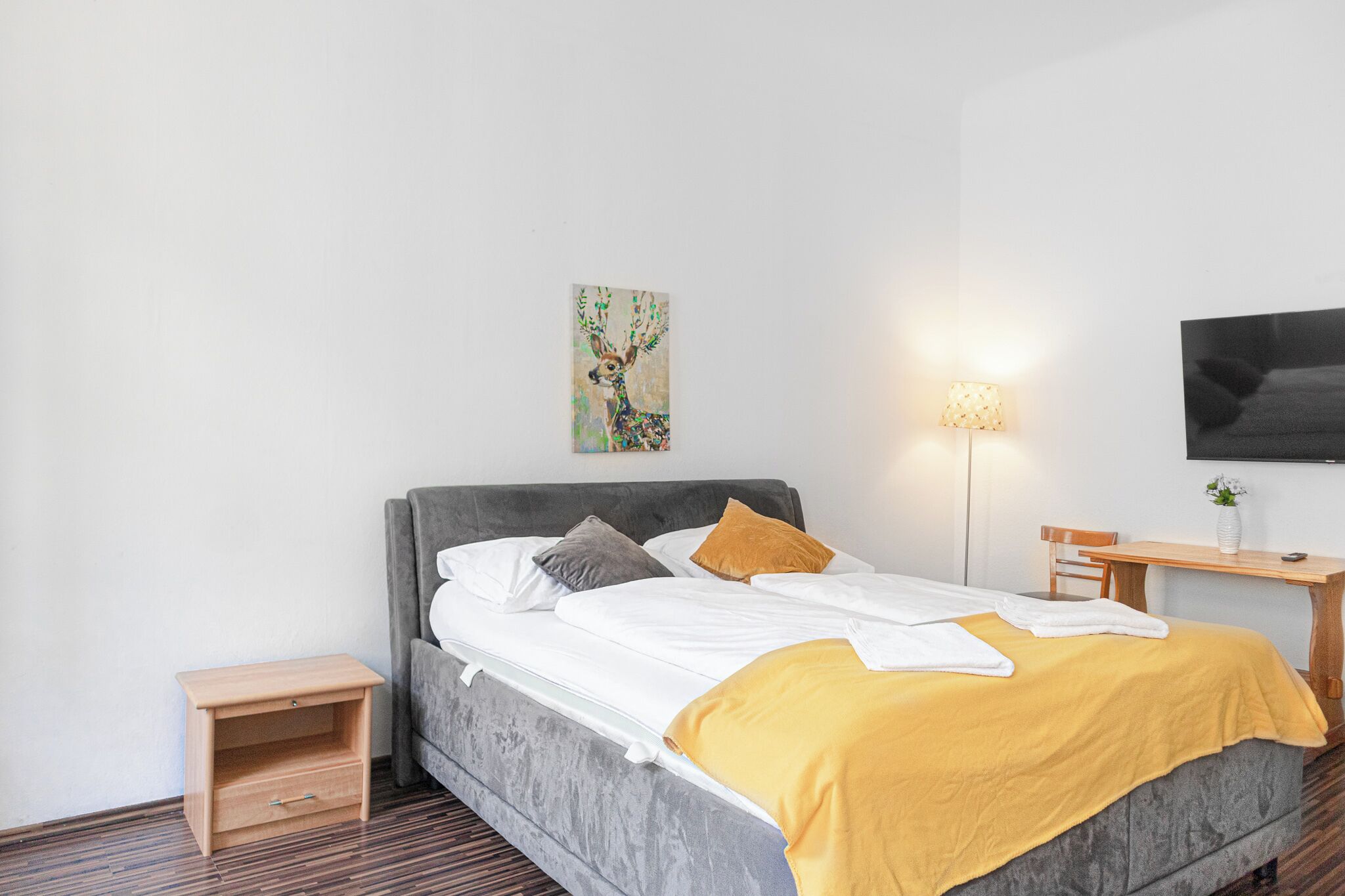 A modern bedroom with a grey upholstered bed, a bedside table, a small wooden nightstand, a bedside lamp, and a wall-mounted TV.