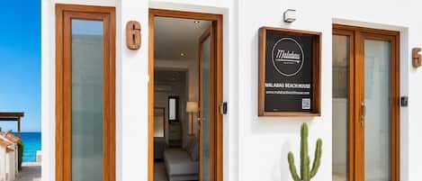 Entrada de hotel con un letrero que dice Malabau Beach House, un cactus en maceta y vista a la playa.