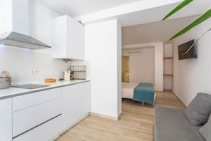 A compact kitchen with white cabinets, a stainless steel range hood, and a small countertop with kitchen appliances. There is a grey sofa and a television mounted on the wall. - 1 beach line. Malabau N4 (Tarifa)