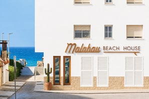 Un hotel frente a la playa llamado 'Malabau Beach House' con vista despejada al mar, un cactus y un camino que conduce a la entrada.