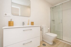 A modern bathroom with a white vanity, a toilet, a shower enclosure, and a mirror. - 1 Linea Playa, Malabau 01 (Tarifa)