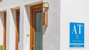 A building facade with three windows and a door, a blue sign with 'AT' and 'CIUDAD' indicating a city location.