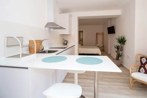 A compact kitchen with a white countertop, two sinks, and a modern stove. A small dining table with two chairs is in the foreground. The room has a neutral color palette and includes a bed and a television in the background.
