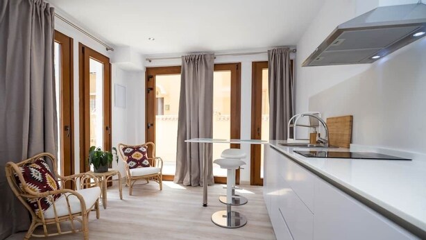A modern kitchen with white cabinets, a stainless steel sink, and a built-in range hood. There are two wicker chairs with cushions and a small table with a plant.