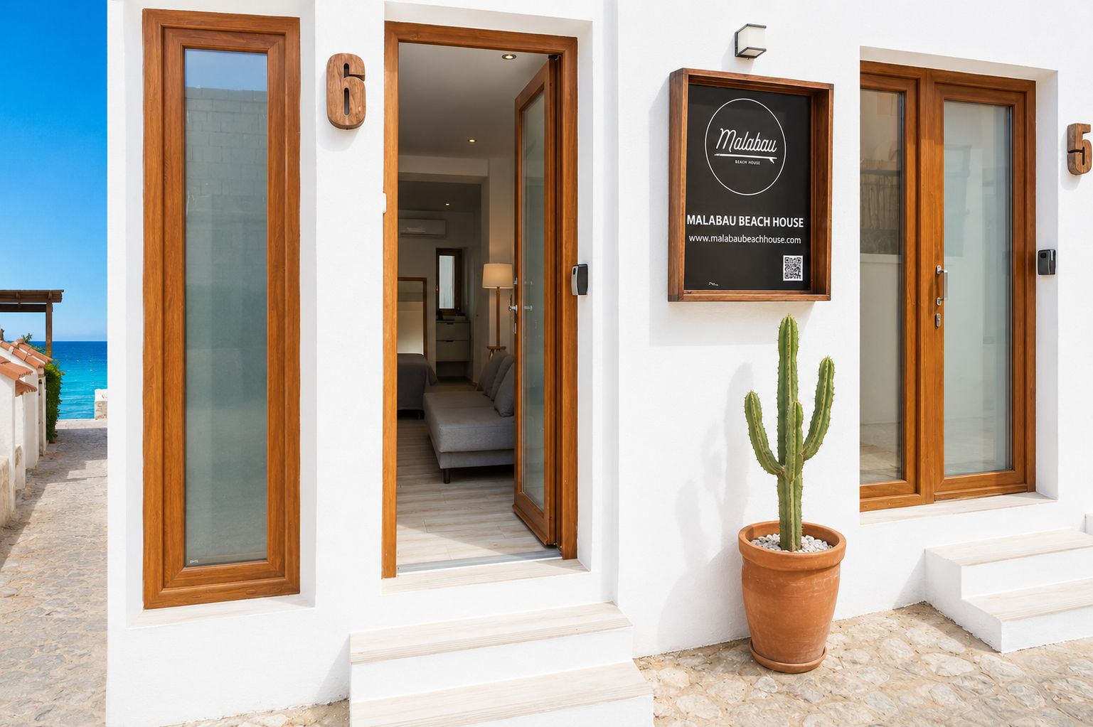 A hotel entrance with a sign for Malabau Beach House, a potted cactus, and a view of the beach.