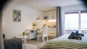 Een slaapkamer met een bed, een eettafeltje, een stoel, een kitchenette en een raam met gordijnen.