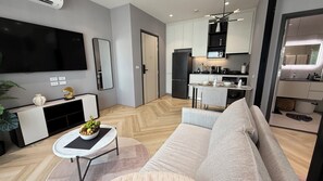 A modern living room with a sofa, a coffee table with fruit, a TV, and a kitchen area with cabinets and appliances. - Elegant Holiday Living at 1 km to Bangtao Beach, 1BR near Bangtao Beach, Phuket (Tambon Choeng Thale)