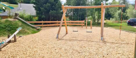 Ein Spielplatz mit Schaukeln und einer Rutsche, umgeben von einem Holzzaun und Bäumen.