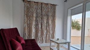A room with a red sofa, a patterned curtain, a small table with a plant, and a view through a glass door. - Djerba 7 Exclusive Stay with Daily Homemade Brunch (Aghir)