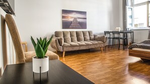 A modern living room with a wooden floor, a grey sofa, a small table with a plant, and a painting of a pier on the wall. - Comfy 2BR Apartment Long Stay Downtown (Vienna)