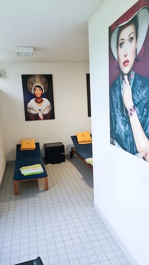 Una habitación con dos camas, un banco, un cuadro en la pared y un pequeño armario negro.