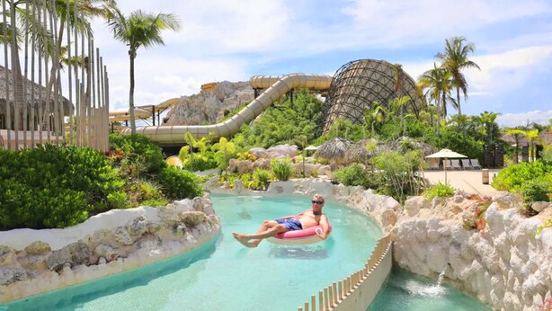 Un hombre flotando en una piscina con un tobogán y formaciones rocosas al fondo.