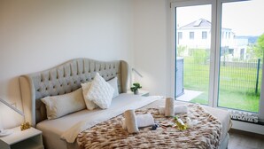 Une chambre avec un grand lit, une table de chevet, une fenêtre donnant sur une maison et une petite plante posée sur la table de chevet.