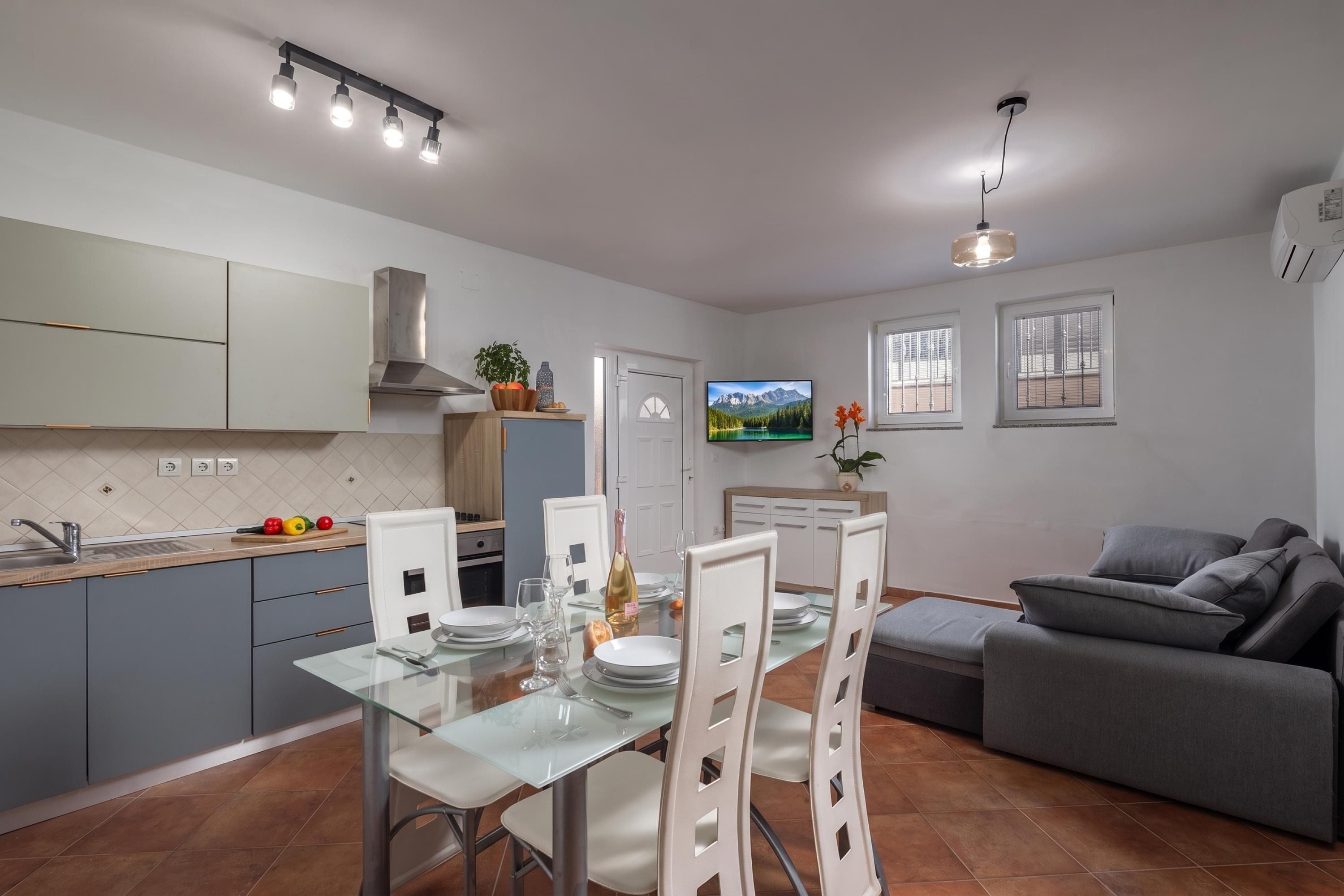A modern kitchen with a dining area, a flat-screen TV mounted on the wall, and a grey sofa in the living room.