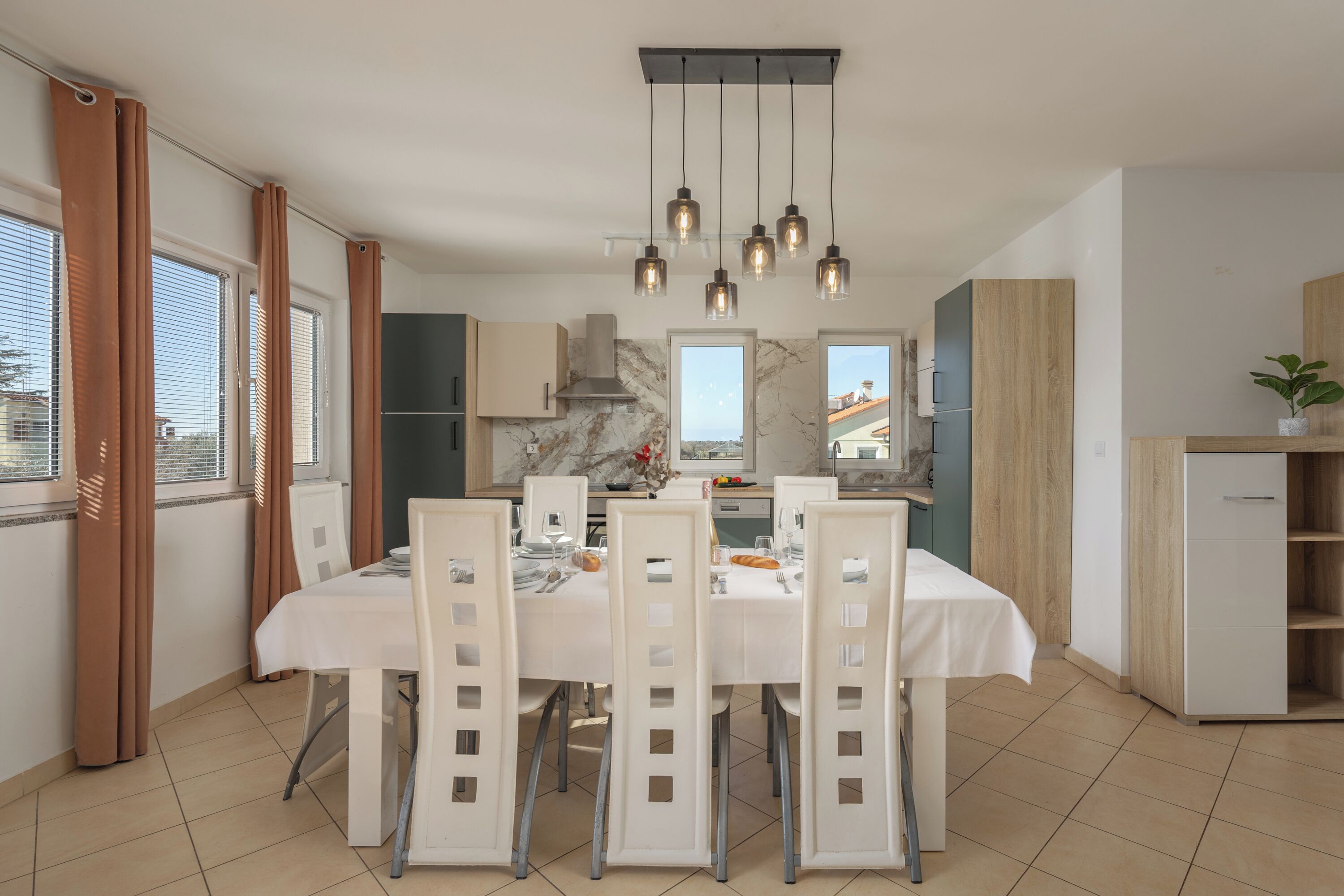A modern kitchen with a dining area, featuring a marble countertop, a built-in stove, and a hanging light fixture.