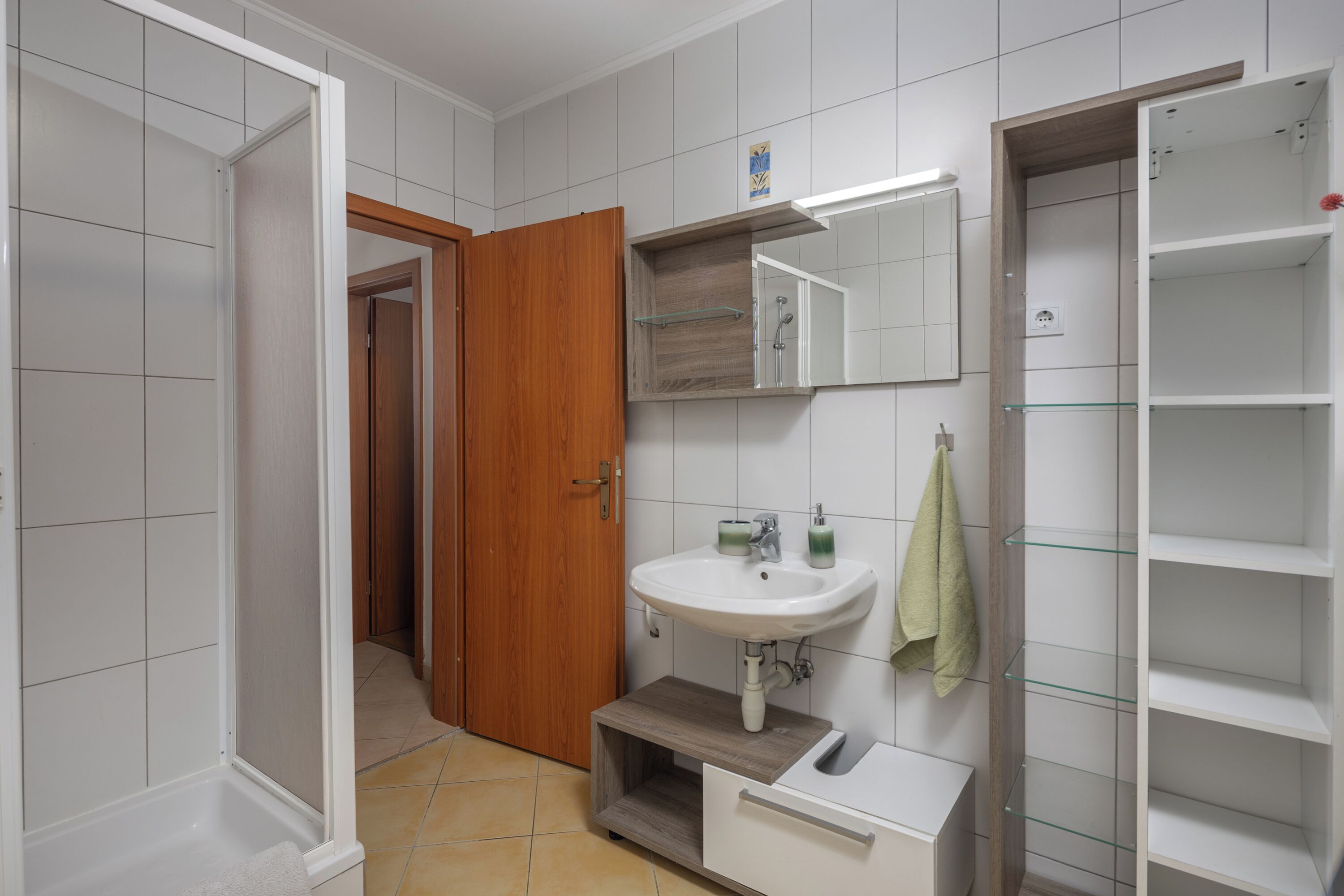 Baño con lavamanos blanco, mueble de madera y estante de vidrio.