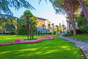 Un jardín bien cuidado con un parterre sinuoso, árboles grandes y un sendero que lleva a un edificio de varios pisos y balcones.