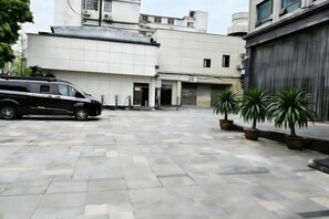 Un patio empedrado con un vehículo negro estacionado a la izquierda, un edificio con garaje a la derecha y varias plantas en macetas.