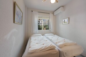 Une chambre avec deux lits simples, une fenêtre avec des rideaux, un appareil de climatisation et un tableau encadré au mur.