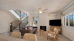 Un salon moderne avec un canapé, des fauteuils en osier, un téléviseur à écran plat, une cheminée et un escalier.