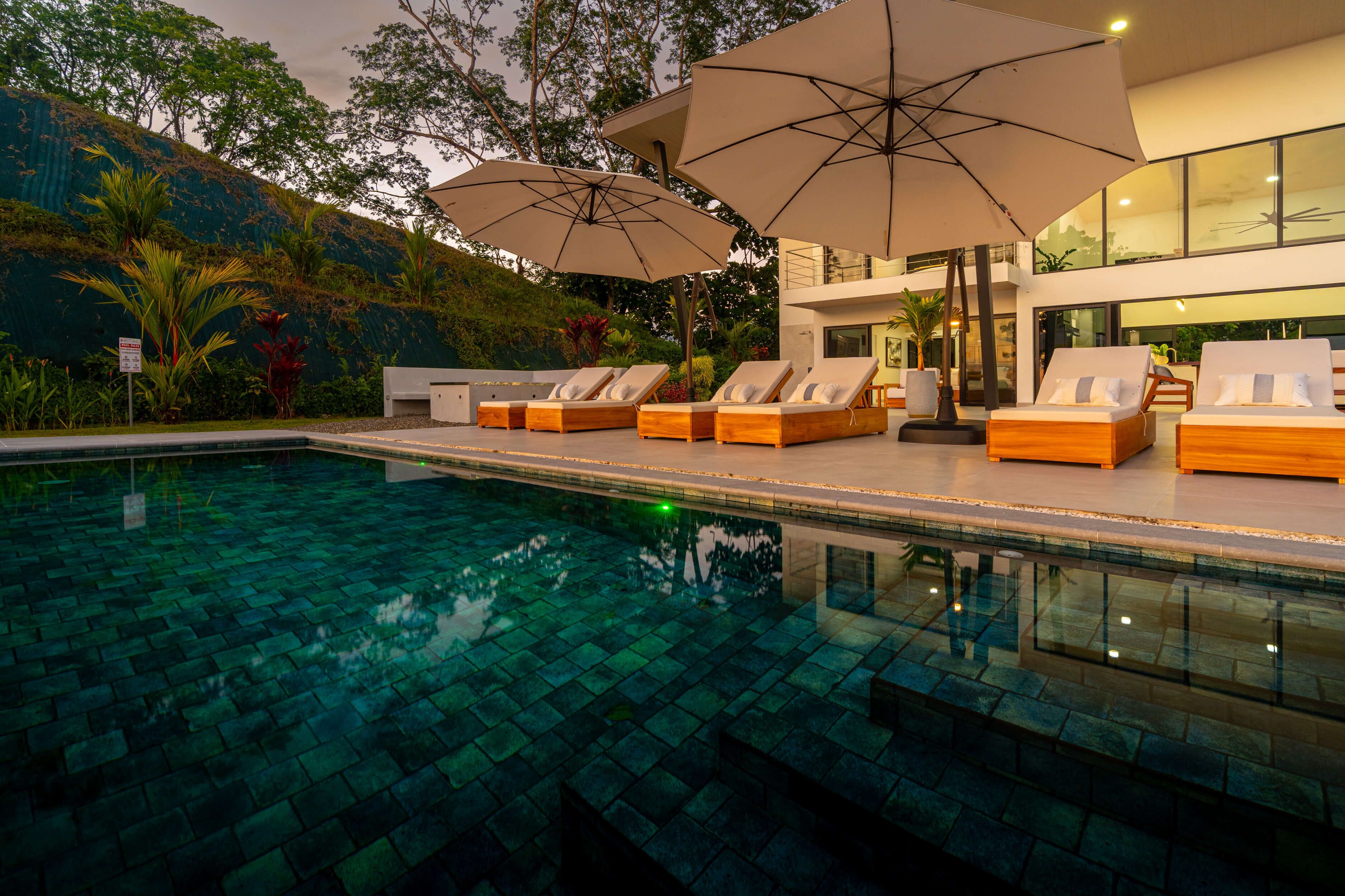 A swimming pool with lounge chairs and umbrellas, surrounded by lush greenery and modern architecture.