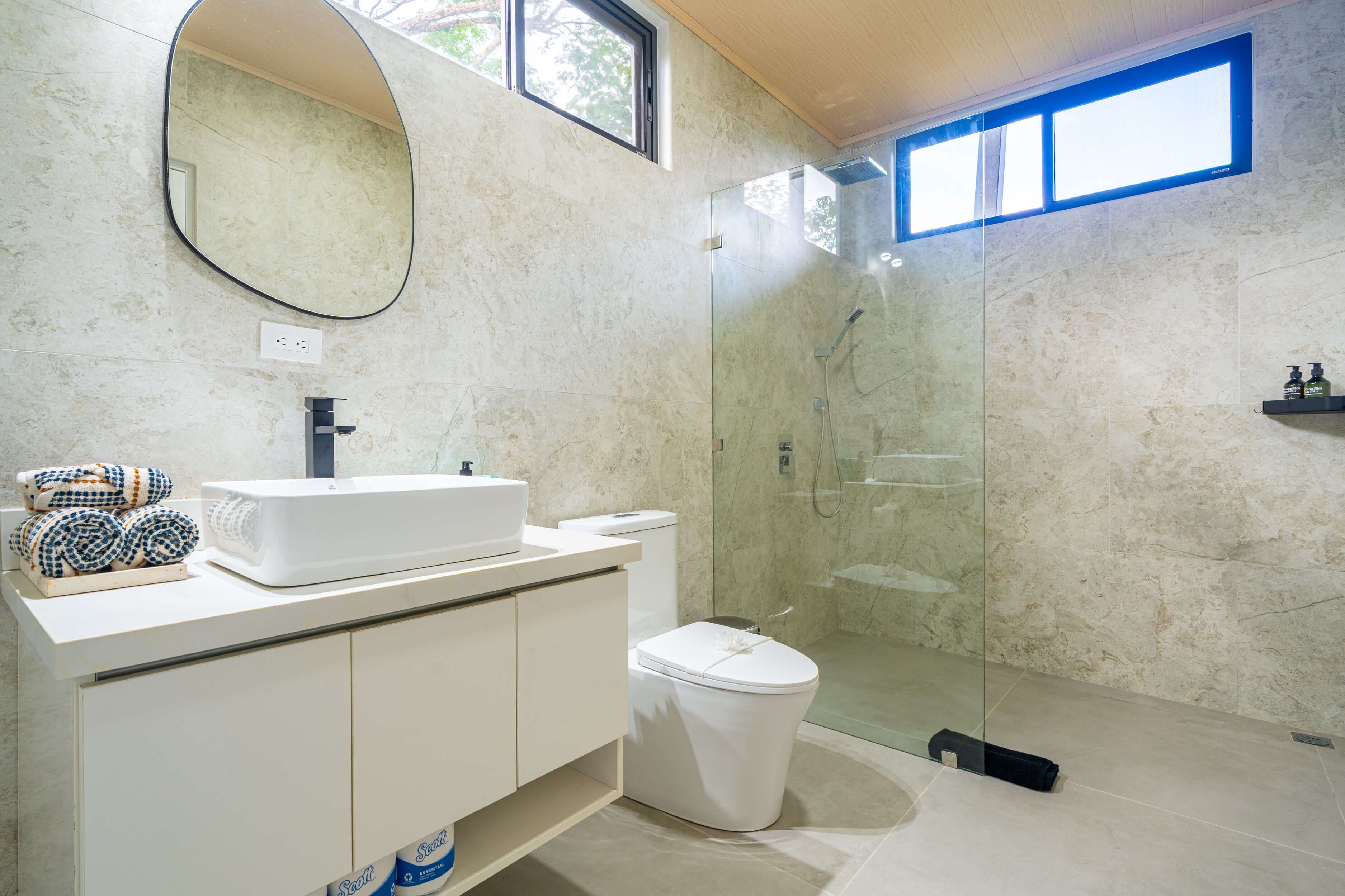 Un baño moderno con un lavabo blanco, espejo y retrete. Cuenta con una cabina de ducha de vidrio y un ventanal pequeño.