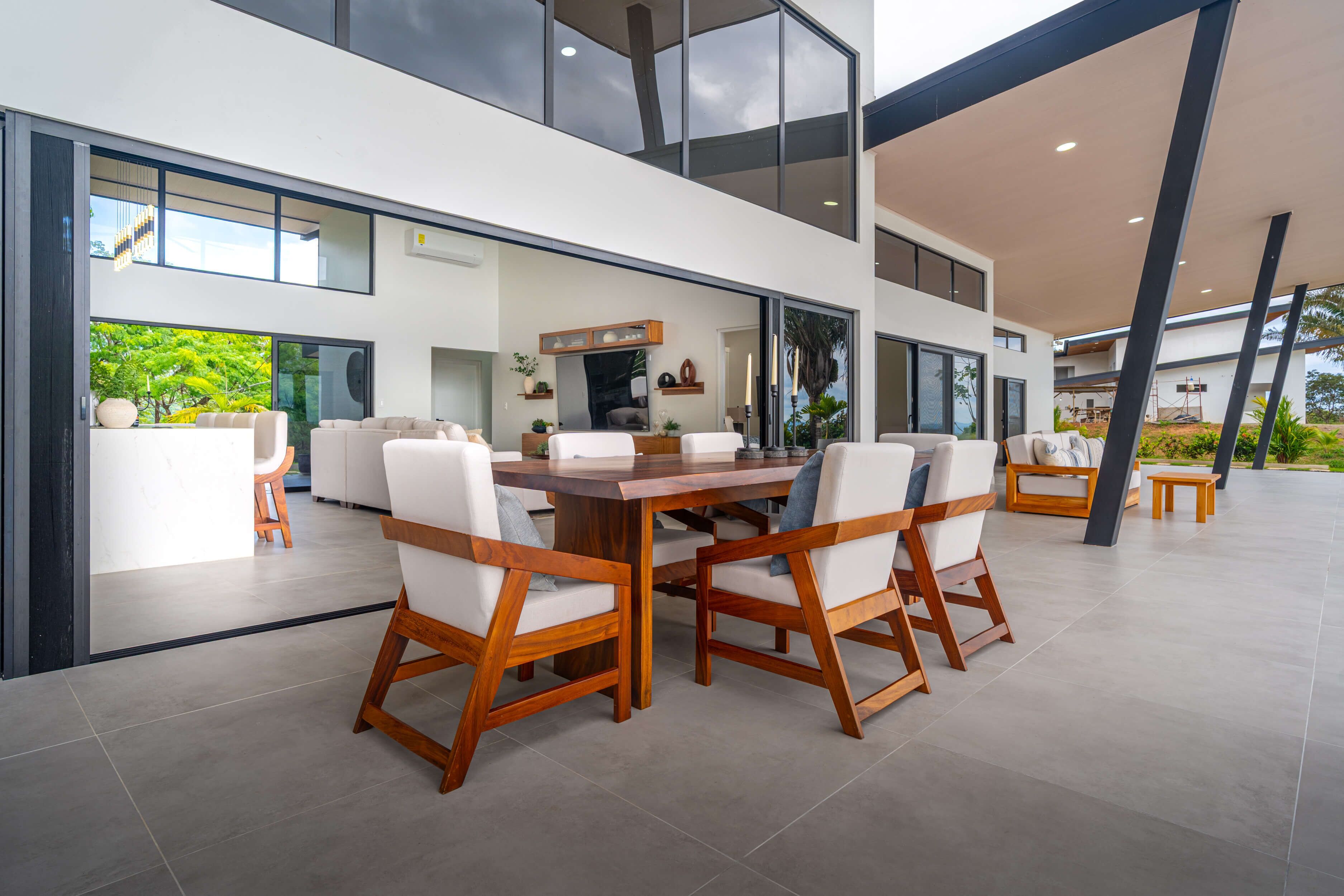 A modern dining area with a wooden table and chairs, large windows, and a view of greenery outside.