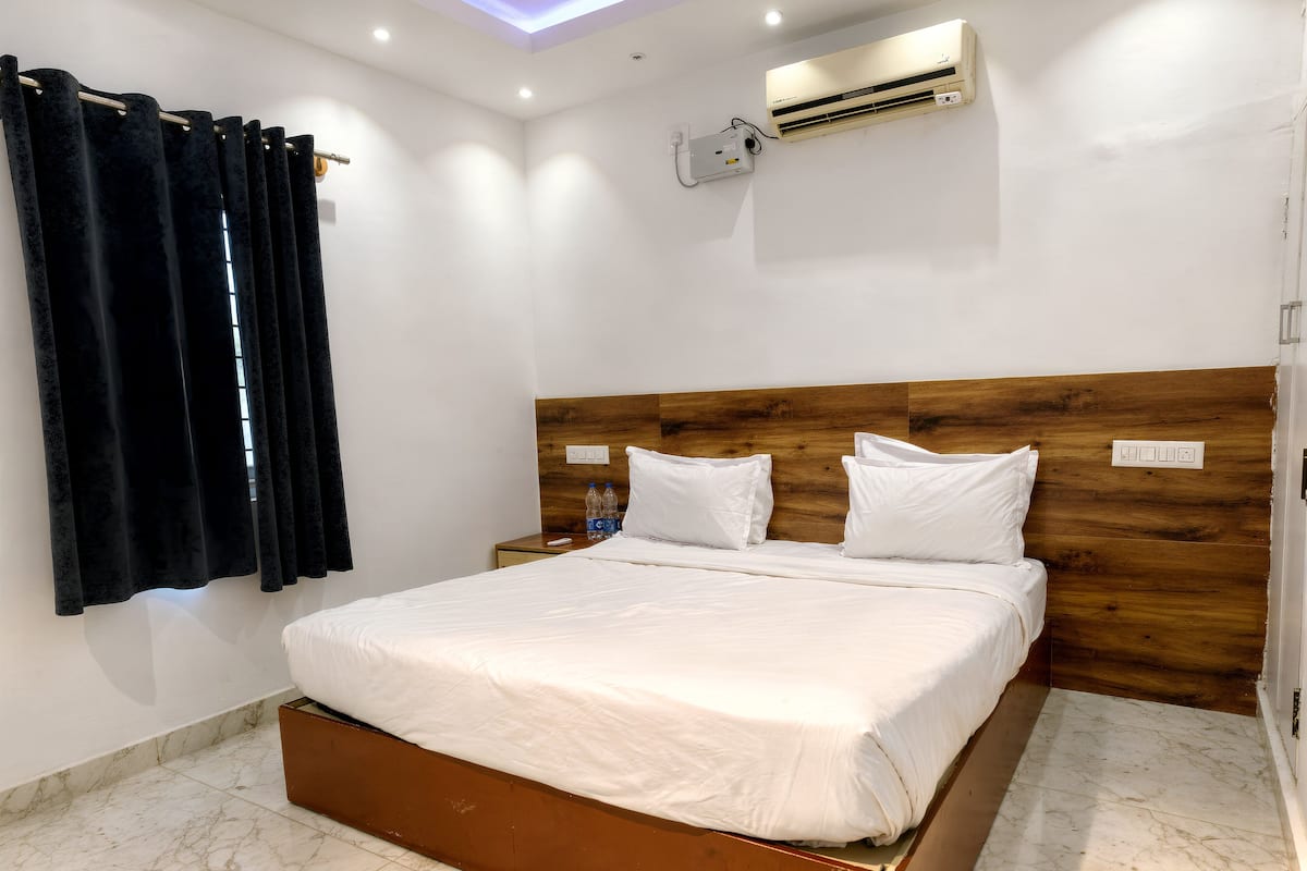 A hotel room with a bed, a wooden headboard, an air conditioning unit, a nightstand, and a window with curtains.