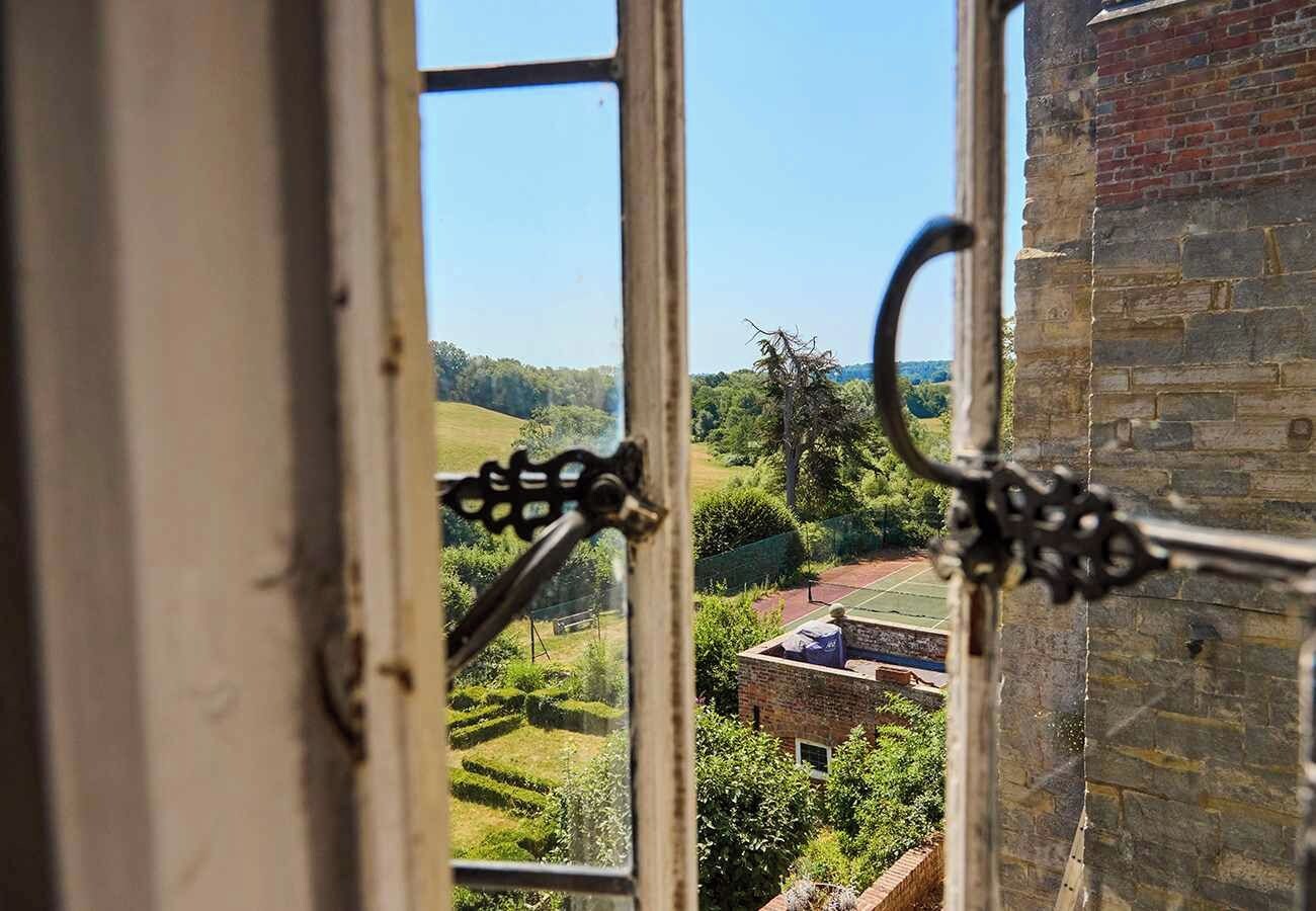 Une vue à travers une fenêtre ornée d’une rambarde en métal, donnant sur un paysage verdoyant avec un bâtiment en briques et un court de tennis.