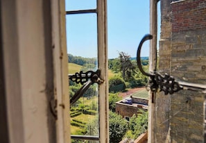 A view through a window with a metal railing, overlooking a green landscape with a brick building and a tennis court.