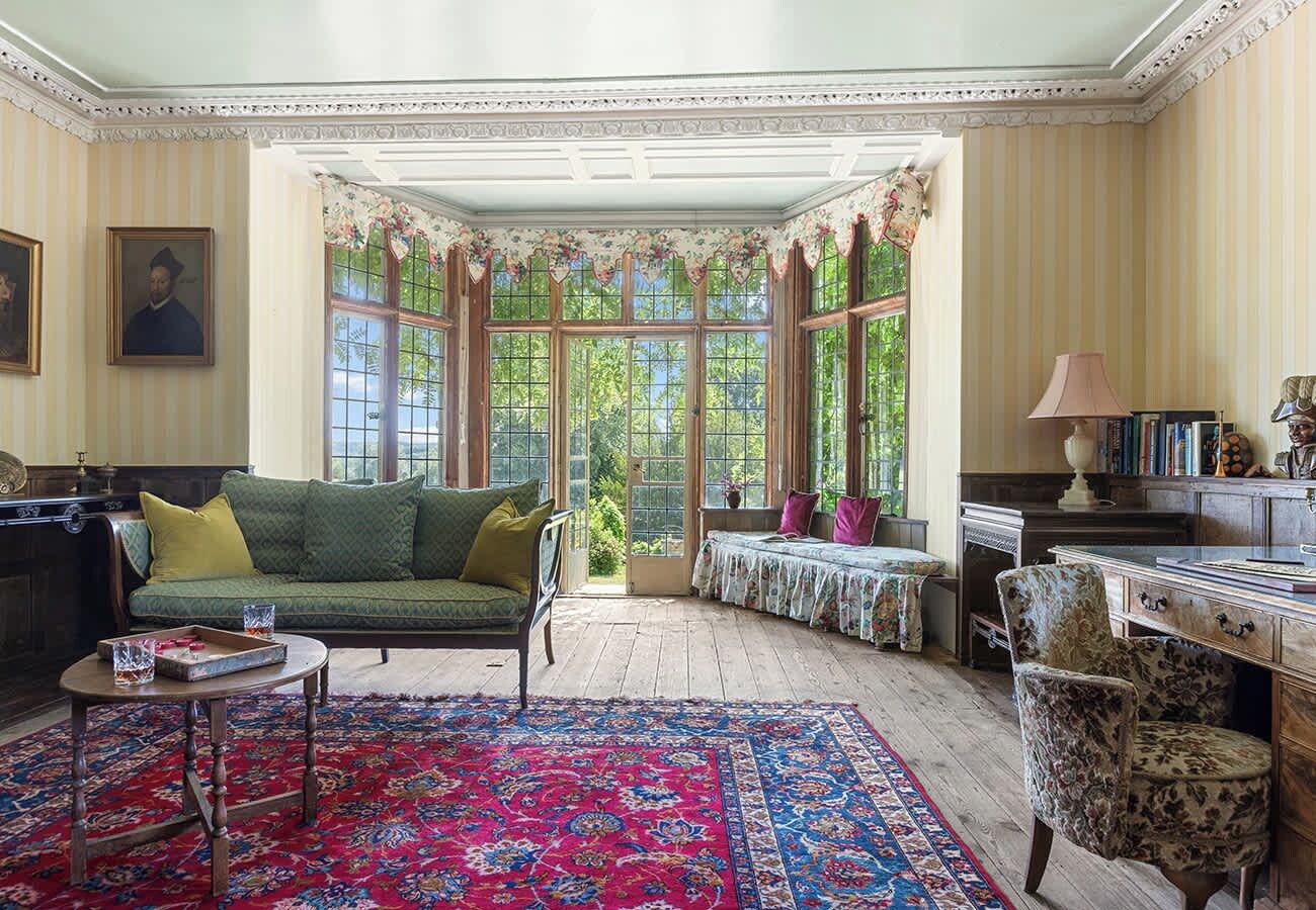 A room with a green sofa, a wooden desk, a patterned rug, and a window with a view of greenery.