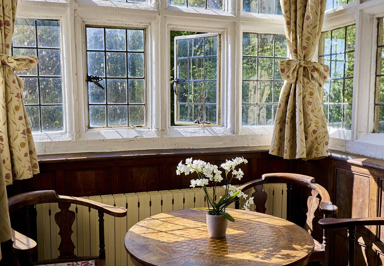 A room with a round wooden table, a potted orchid, and a window with curtains tied back.