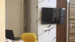 A room with a marble wall, a mounted flat-screen TV, a yellow chair, and a desk with a laptop. - Niharika Heights Luxury Hotel (Bengaluru)