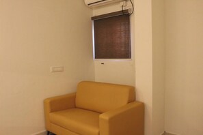 Ein Eckzimmer mit einem einzelnen gelben Sessel, einem Fenster mit brauner Jalousie und einer Wand-Klimaanlage.