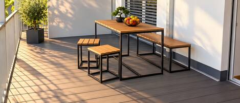 Una terraza de madera con una mesa y bancos, rodeada de muros y un ventanal con persianas.