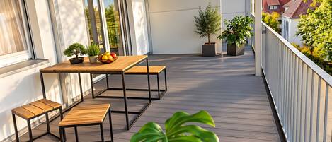 Een balkon met een houten tafel en bankjes, bloempotten en uitzicht op de omliggende gebouwen.