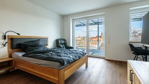 A modern bedroom with a wooden bed, a black chair, a desk, and a large window with a city view. - Premium Apartment City View 8 with XXL Balcony (Pfullendorf)