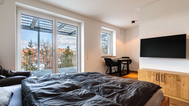 A bedroom with a bed, a desk, and a TV mounted on the wall. - Premium Apartment City View 8 with XXL Balcony (Pfullendorf)