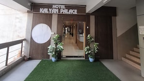 L’entrée d’un hôtel, avec une enseigne indiquant « Hôtel Kalyan Palace », un comptoir d’accueil, un escalier et des plantes en pot.