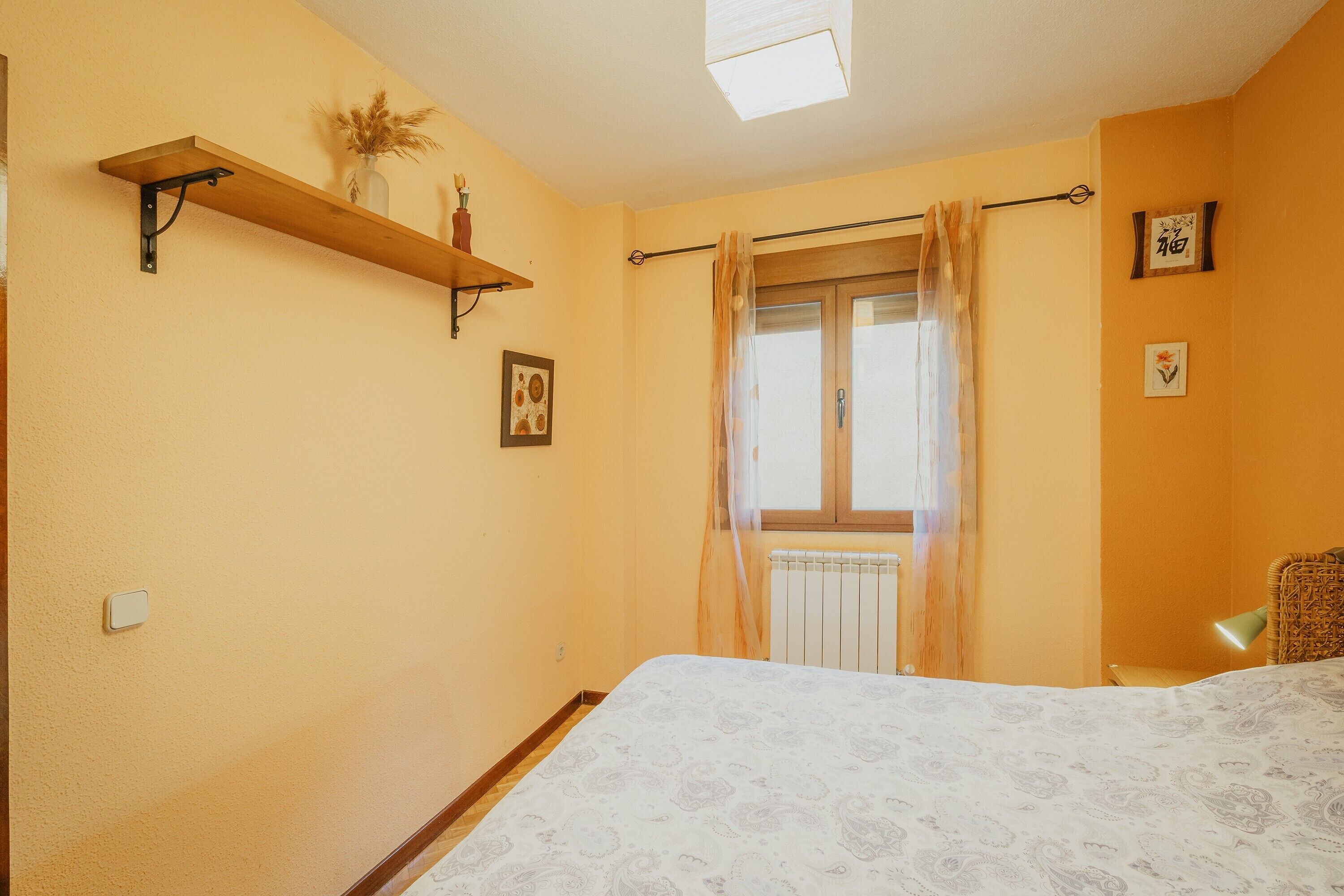 Un dormitorio con cama, ventana con cortinas, radiador, estante con un jarrón y un cuadro en la pared.