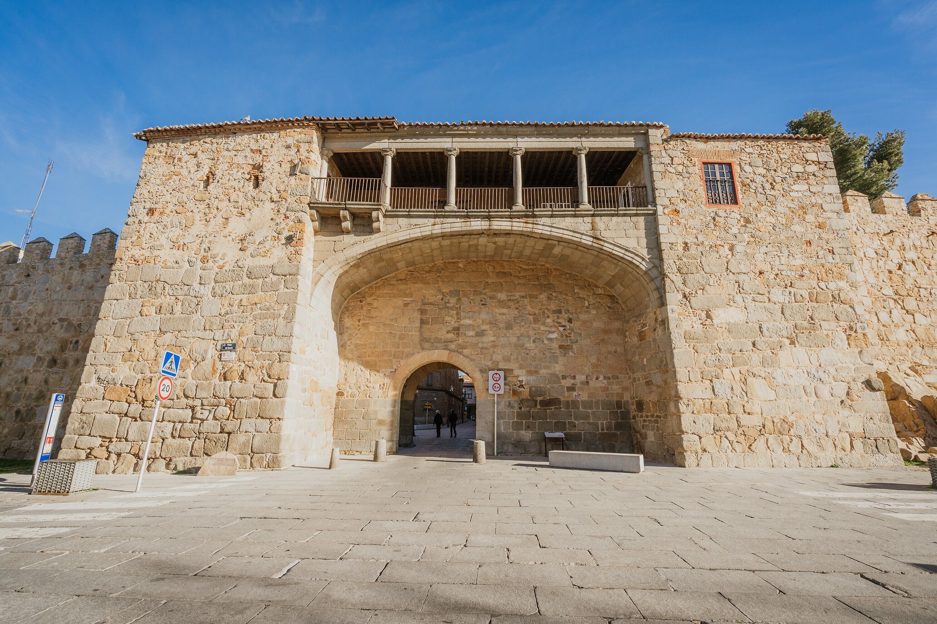 A stone archway leading into a building with a balcony and a sign indicating a 20 km/h speed limit.