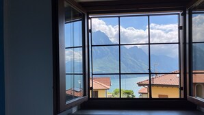 A room with a large window offering a view of mountains and a lake. - Corvino Lake Apartment (Riva di Solto)